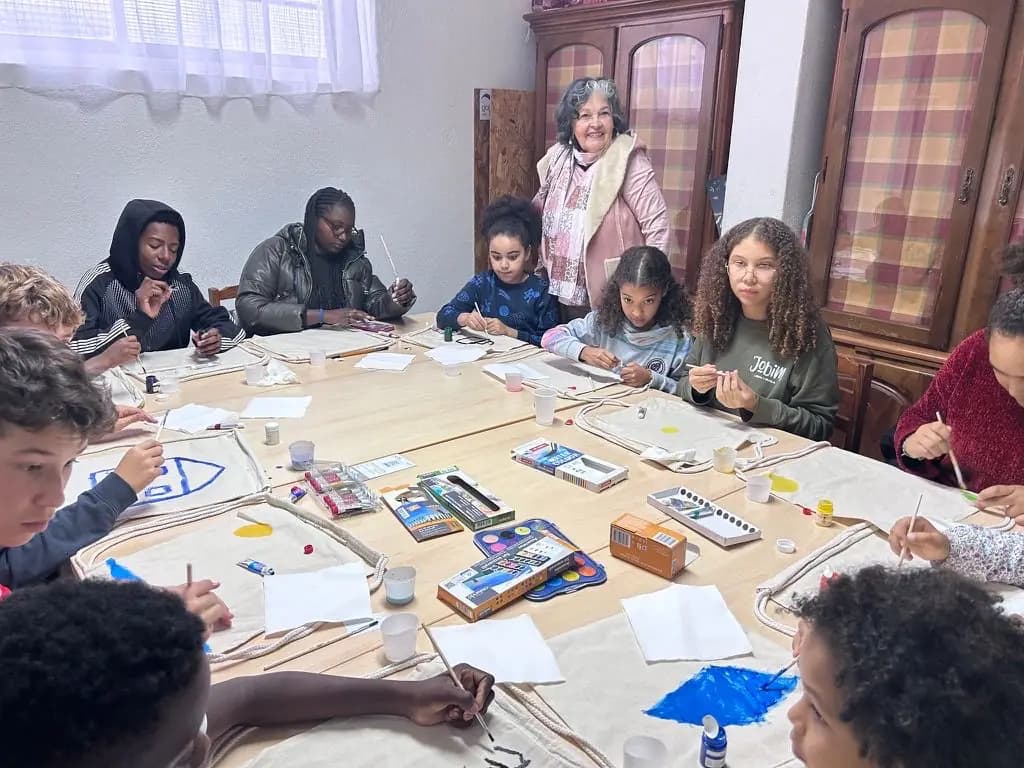 Crianças à volta da mesa grande, a pintar as suas mochilas de pano
